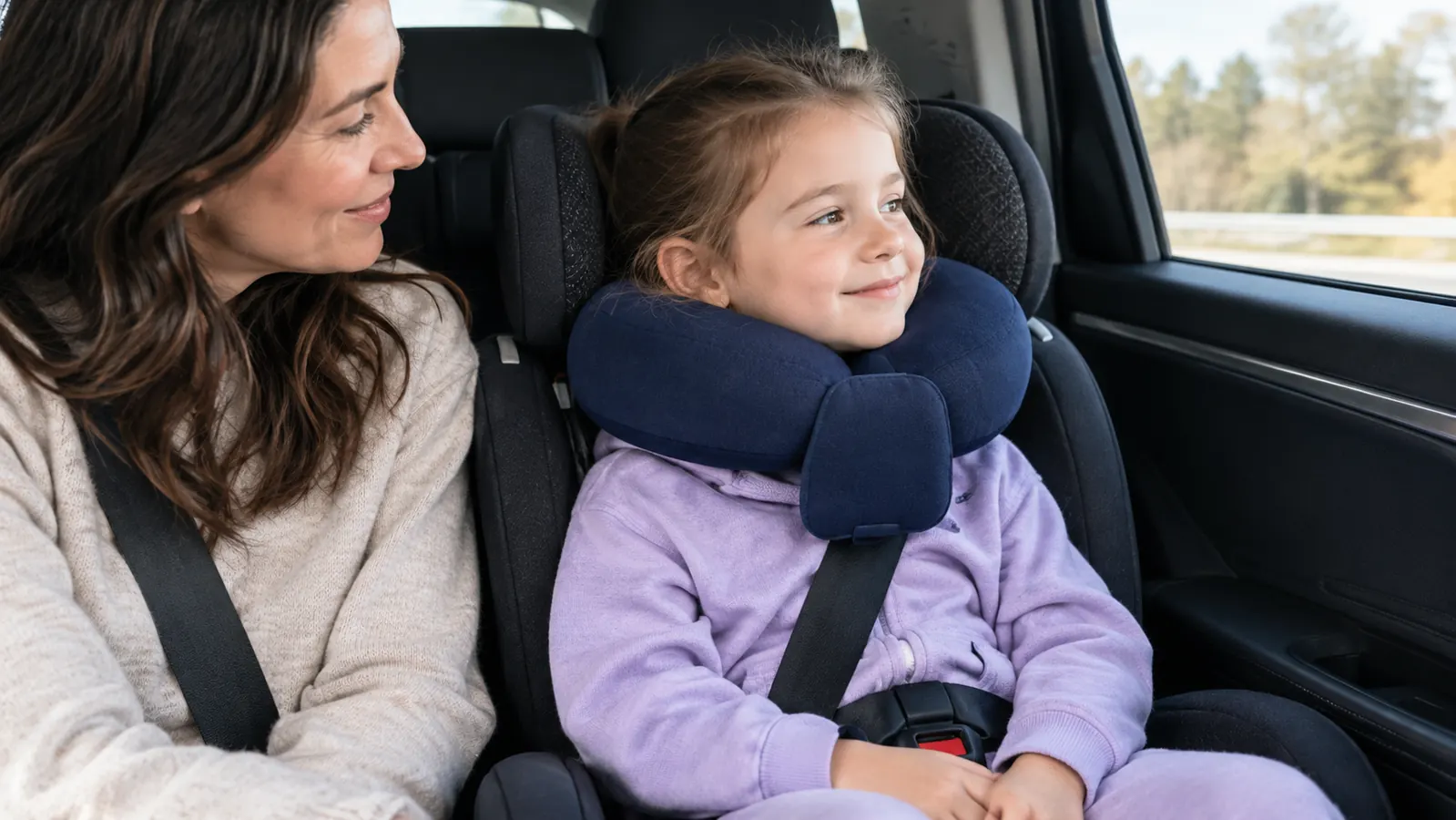 A child wearing a wrap-around travel pillow in a car seat, chin resting on the front support, looking comfortable