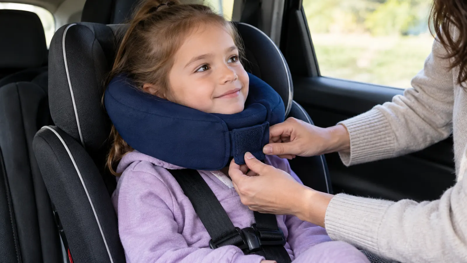 A child wearing a wrap-around travel pillow in a car seat, chin resting in the front support
