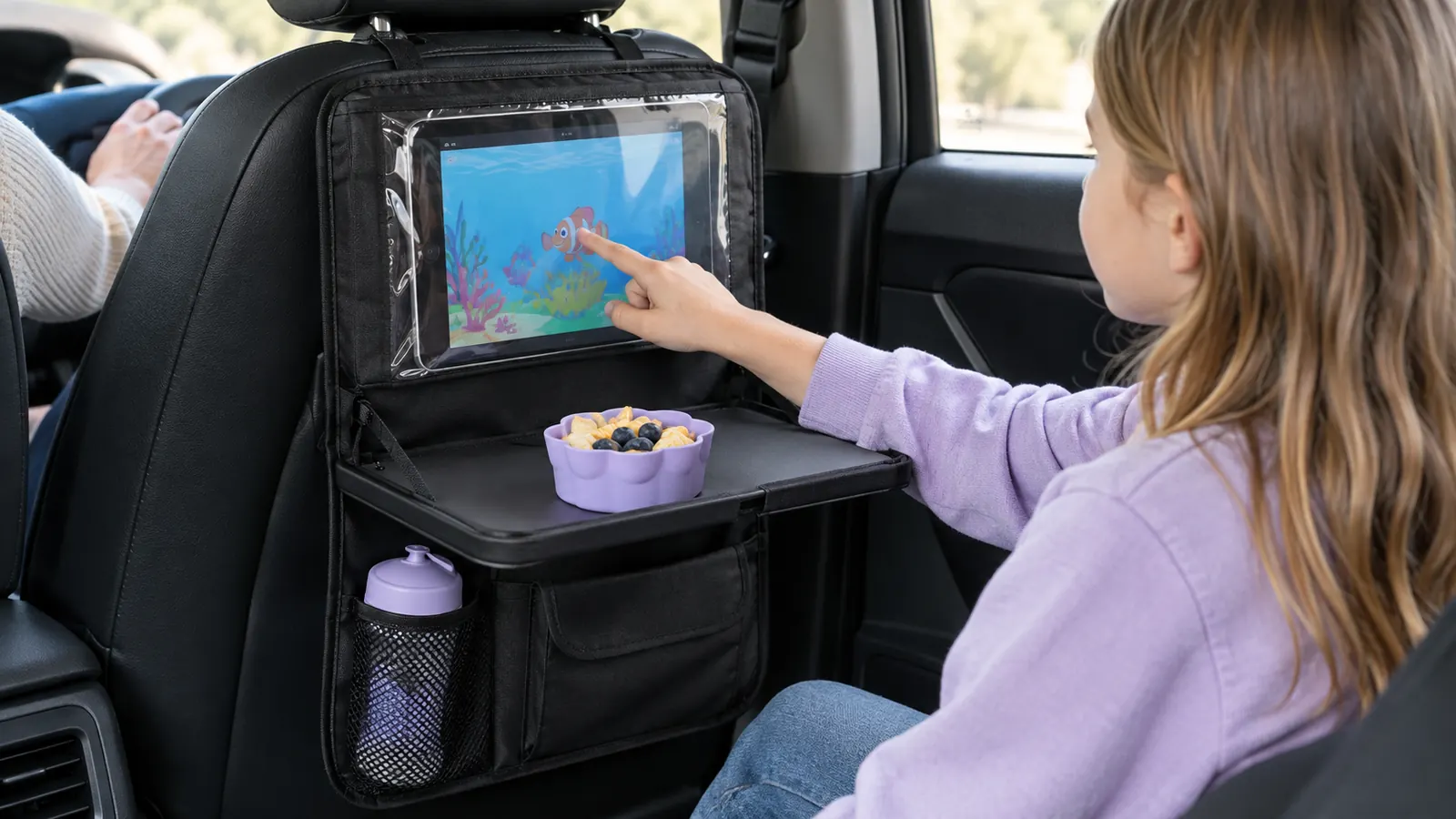 Child's hand using a tablet in the clear pocket of a backseat organizer, with a snack cup on the fold-down tray below it