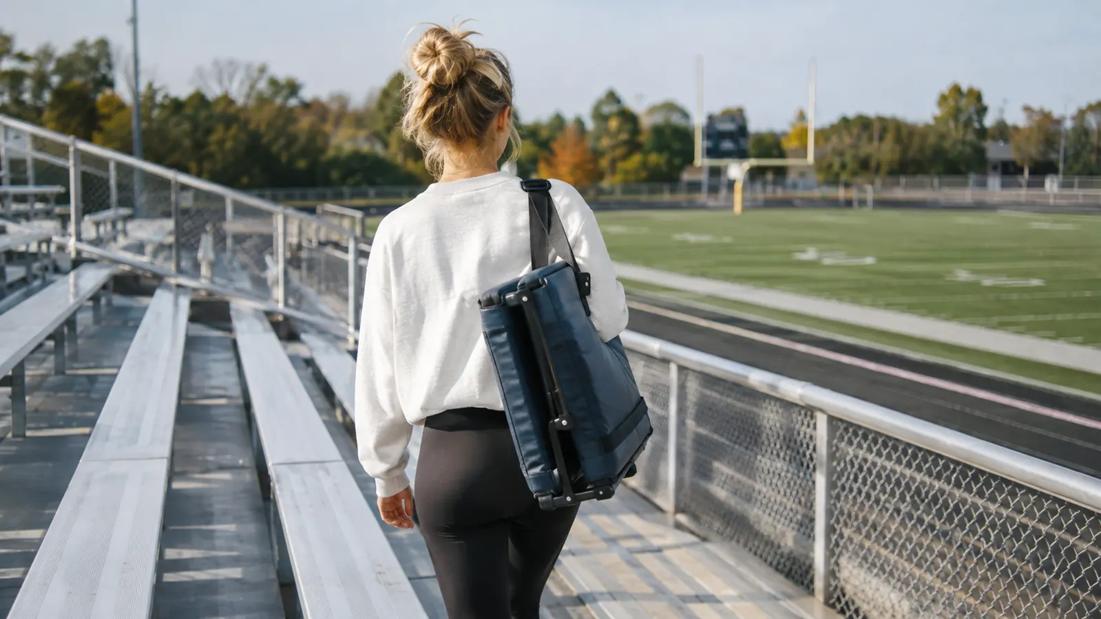 A padded stadium seat folded flat and carried over a shoulder by its strap at a football field