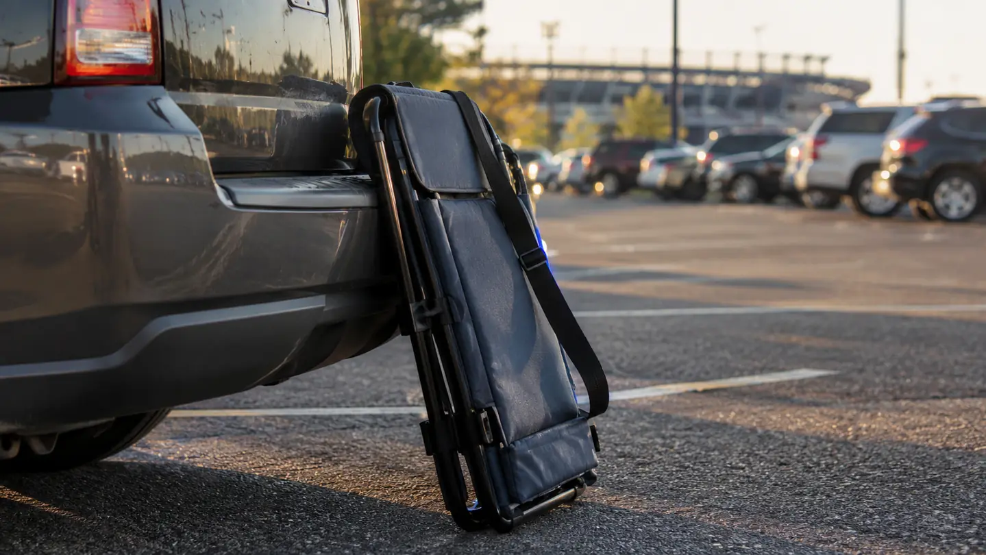 A folded padded stadium seat with a shoulder strap leaning against the bumper of a minivan in a stadium parking lot