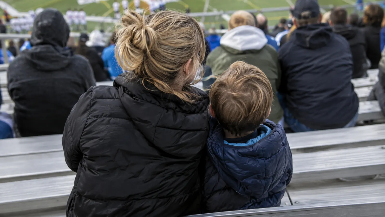 A parent and child sitting on a stadium seat on a metal bleacher, seen from behind at a distance
