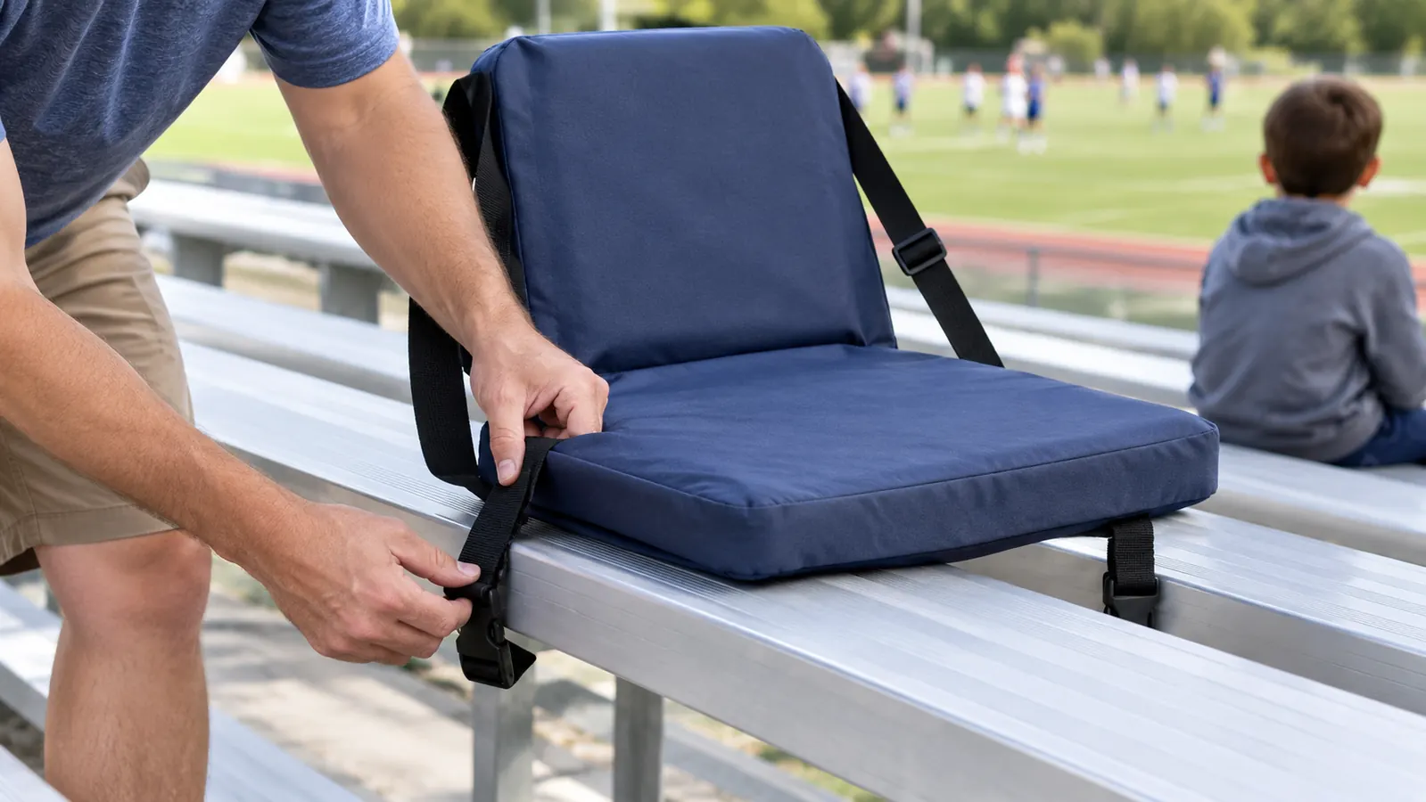A parent's hands unfolding a padded stadium seat and clipping it onto a bleacher bench before a game