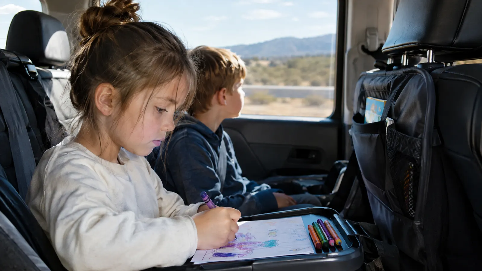 Two children in the back seat of a minivan, one coloring on the fold-down tray of a backseat organizer while the other watches